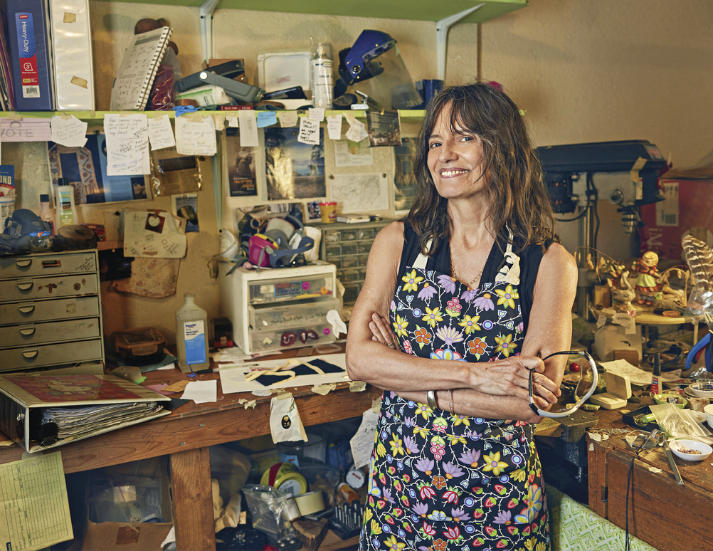 Image of Big Knife in her studio in Oklahoma. She is looking at the camera and smiling while she is surrounded with lots of supplies. She is wearing a black shirt with a colorful apron full of flowers. 