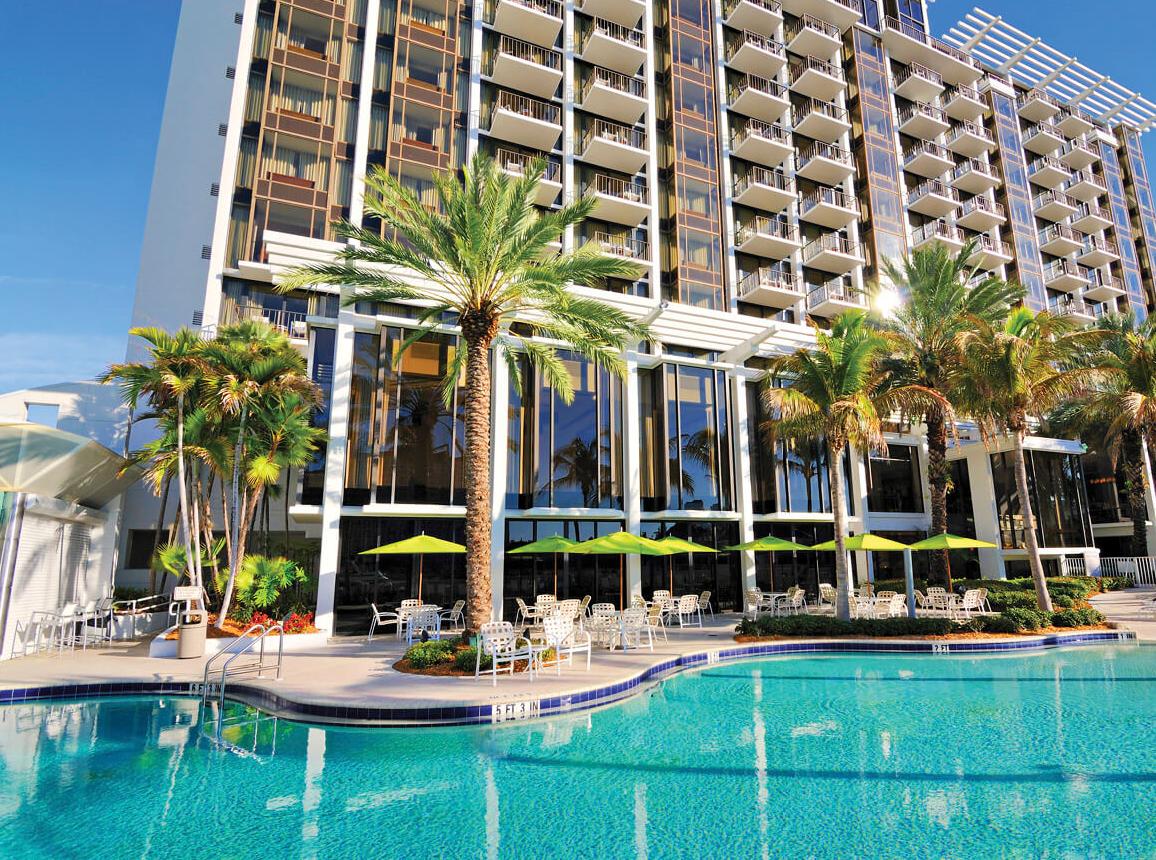 Photograph of a big hotel with a pool and coconut trees in front of it 