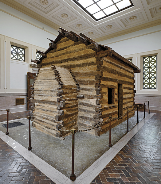 A rustic, one-room log cabin is preserved inside a modern museum gallery with high ceilings and polished floors.