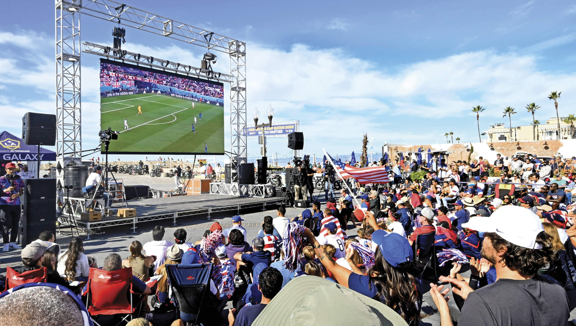 Photograph of a World Cup watch party happening in California. There is a bod crowd of people sitting outside in foldable chairs watching the match play on a big screen in the middle of them 