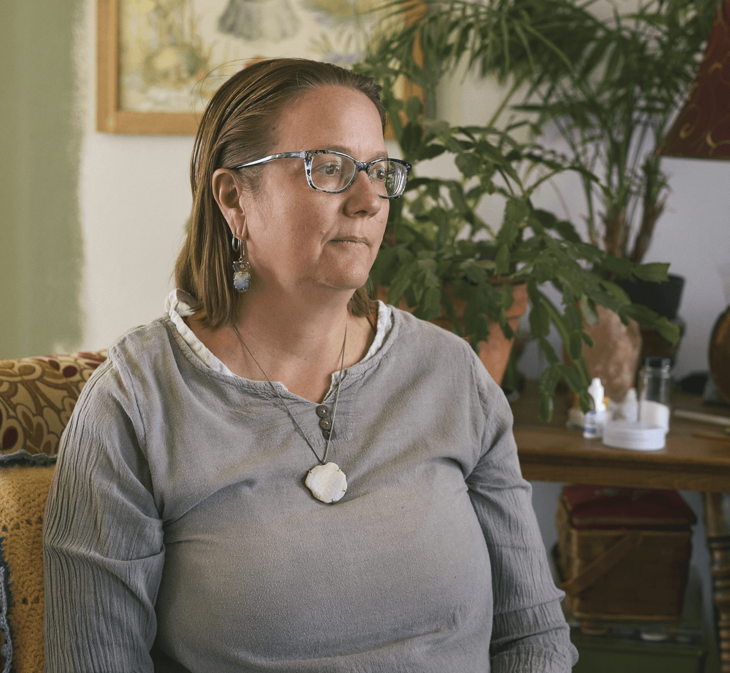 Photograph of Rangel sitting in a living room. She is wearing a grey long sleeve top and a long chain with a ceramic pendant 