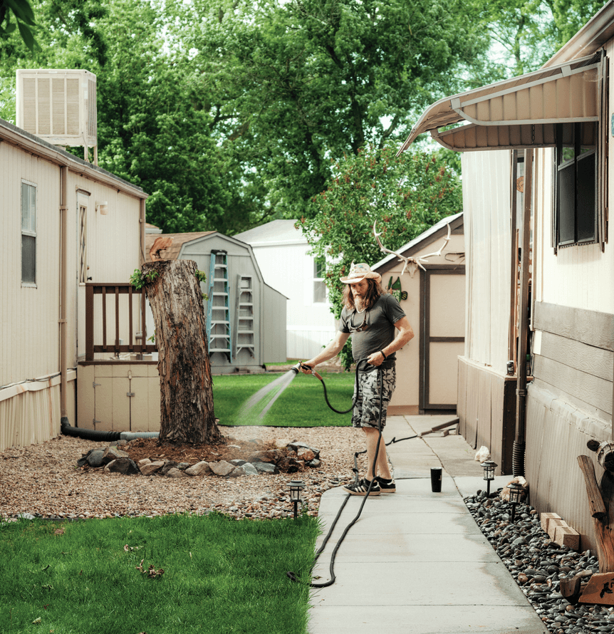 Photograph of Jimmy Krecklow watering the plants of the next-door property. He is wearing shorts and a cowboy hat 