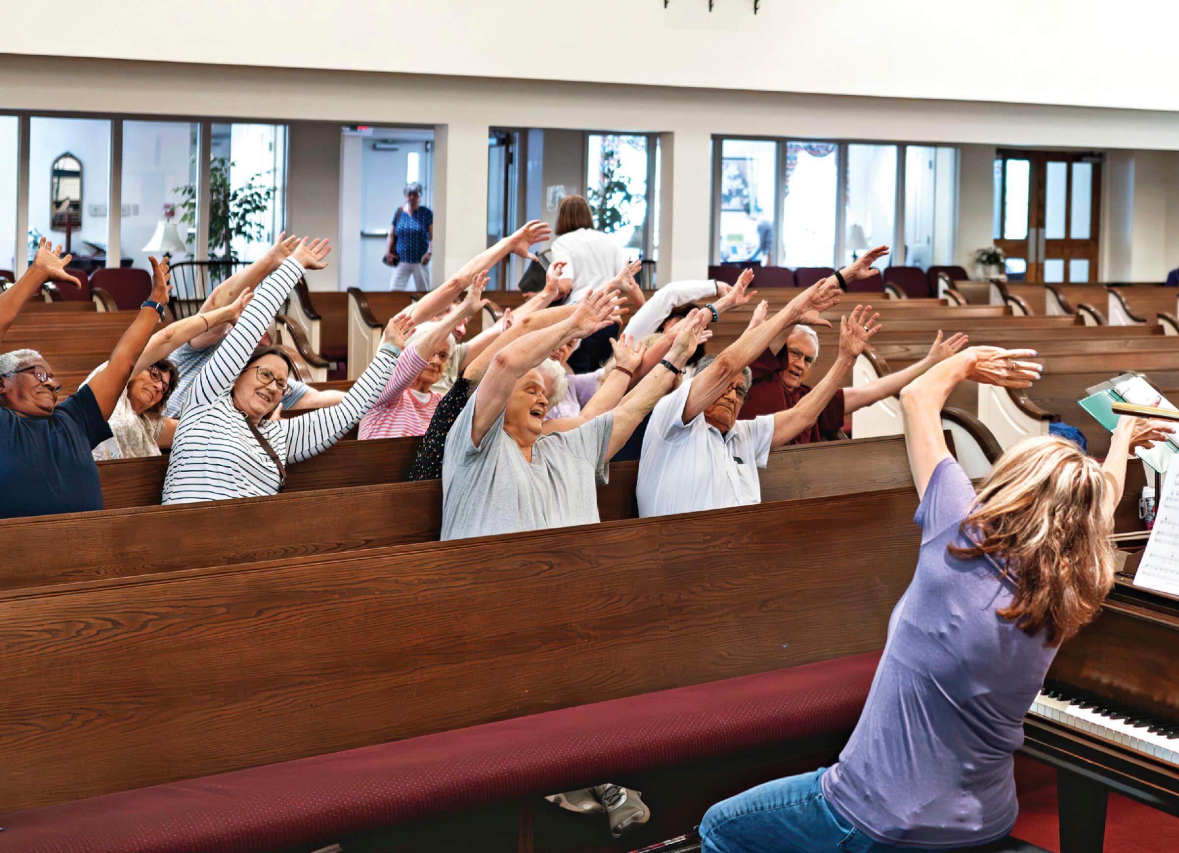 Photograph of Susan Dommer sitting at the front of a church, facing a crowd of people singing and waving their hands in the air and singing together 