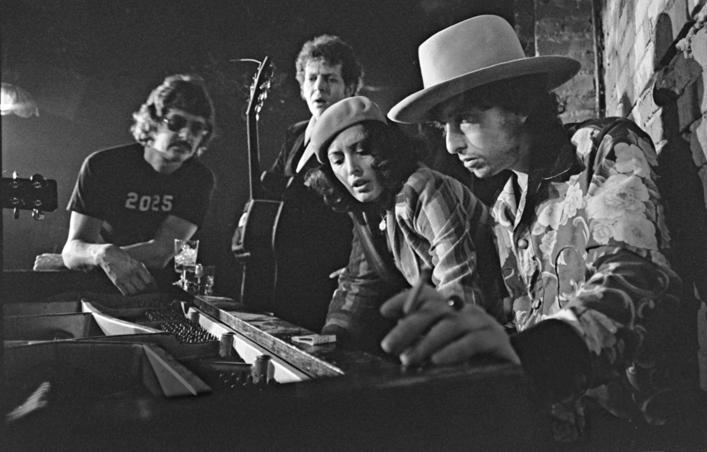 Black-and-white photo of Bob Dylan at the piano with Bobby Neuwirth, David Blue and Ronee Blakley