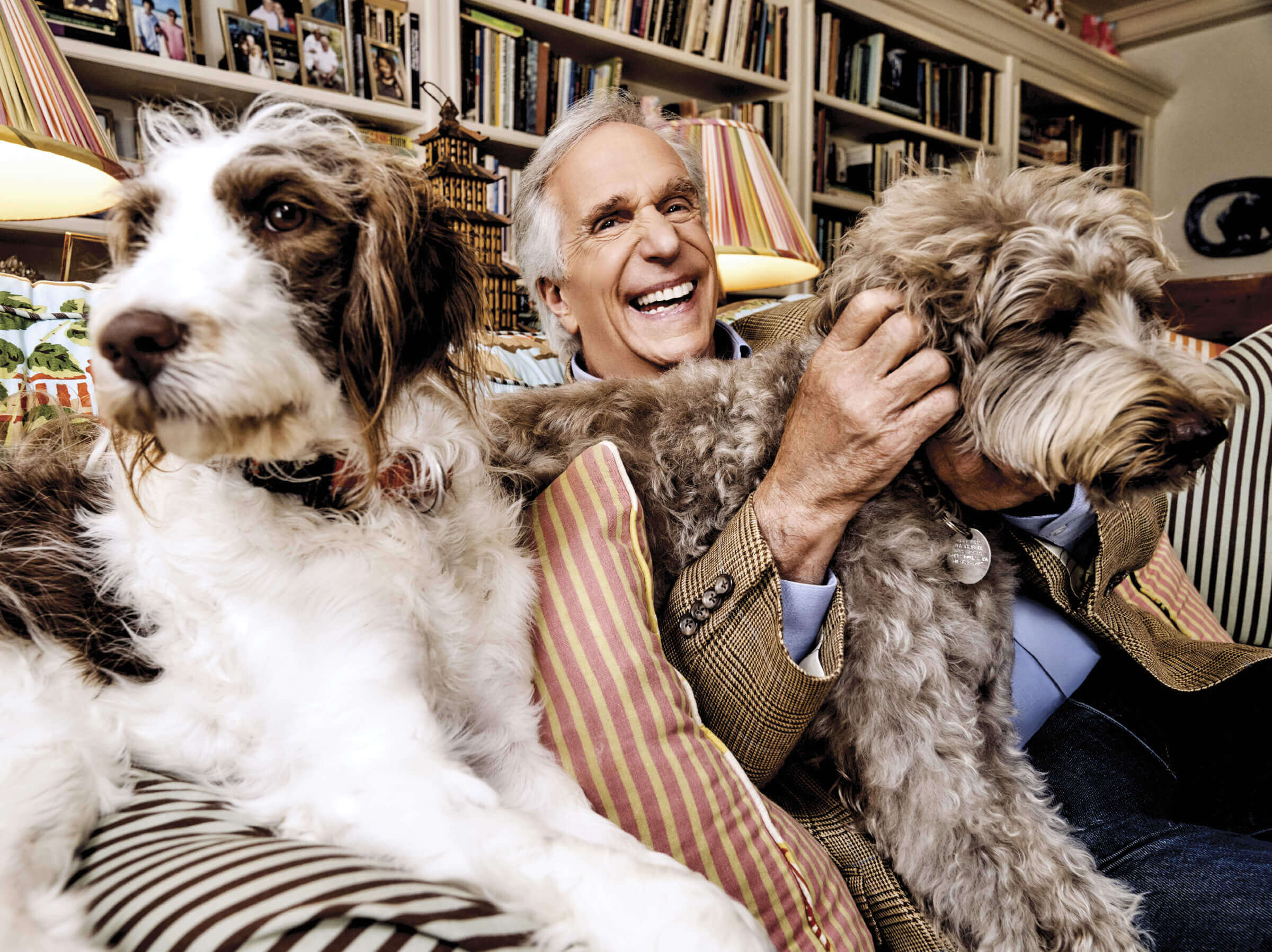 Photo of a laughing Henry Winkler sitting on a couch between his two dogs