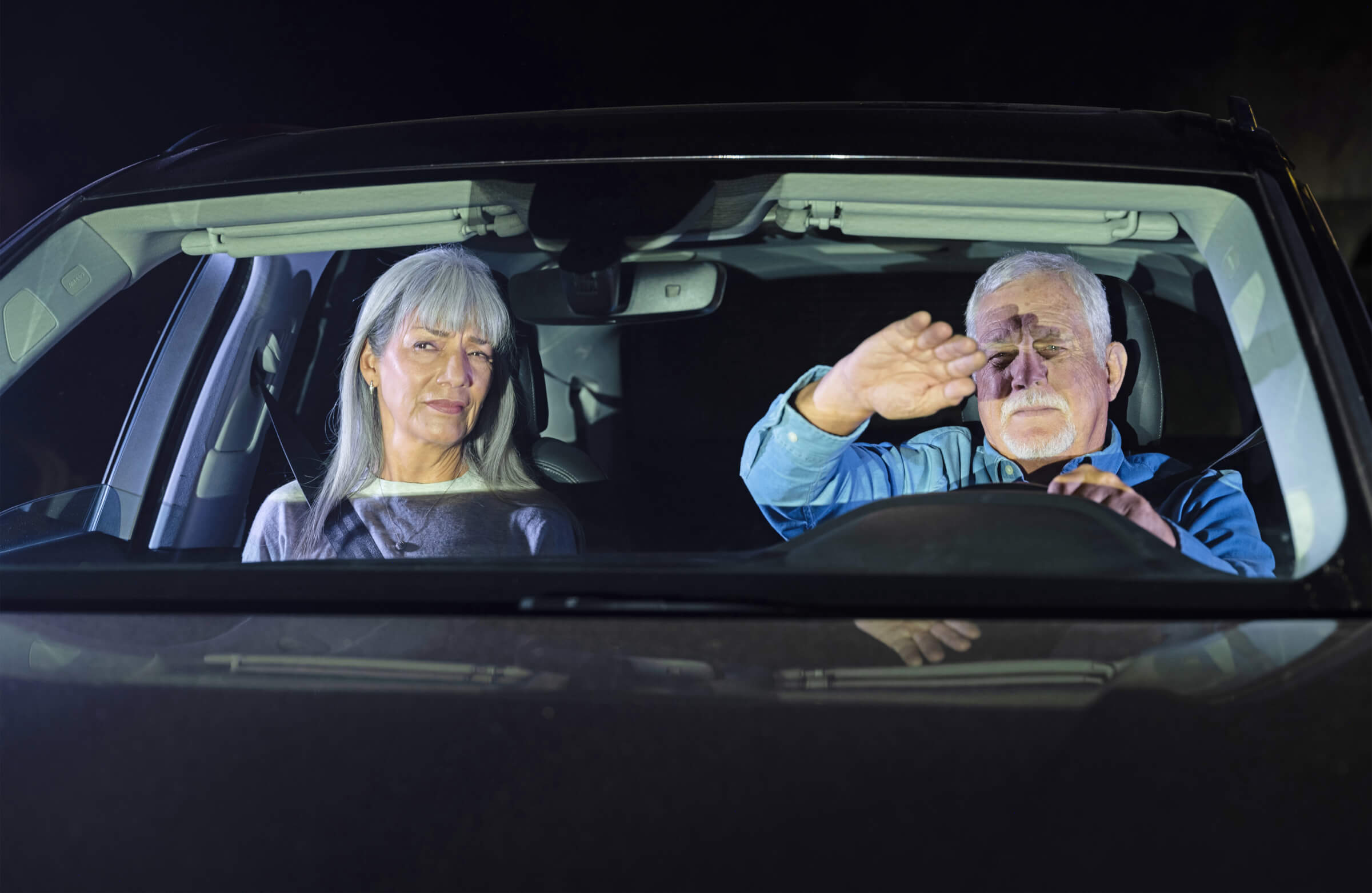 Photo of a couple driving a car at night. The male driver is holding his hand up to shield his eyes from oncoming bright lights.