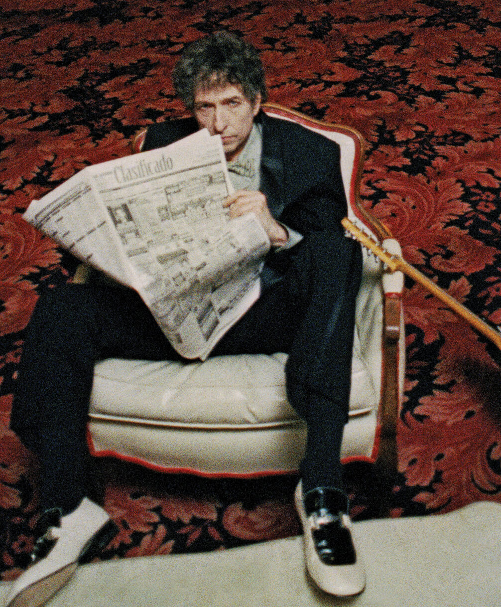Photo of Bob Dylan sitting in a large white easy chair holding an open newspaper in 1999