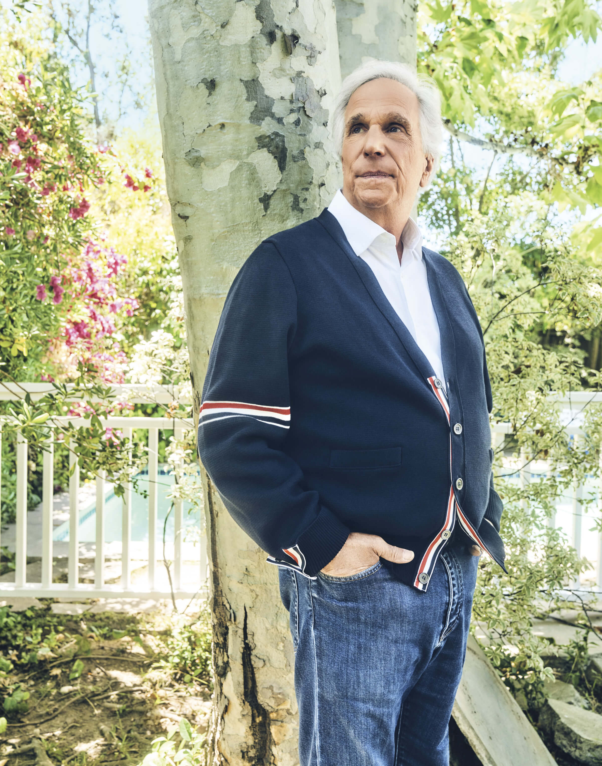 Photo of Henry Winkler wearing blue jeans and a navy blue sweater standing outside near a tree