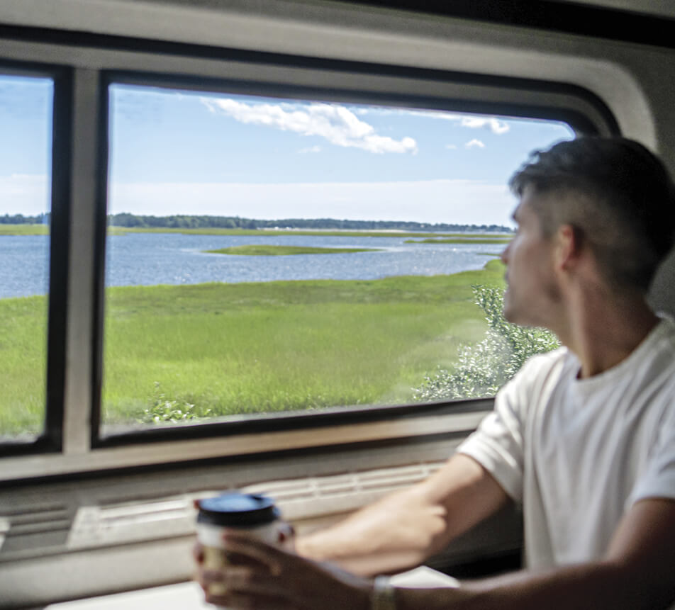 A man enjoying a cup of coffee while looking out the window of a train to see grasslands and water on the Amtrak Downeaster train