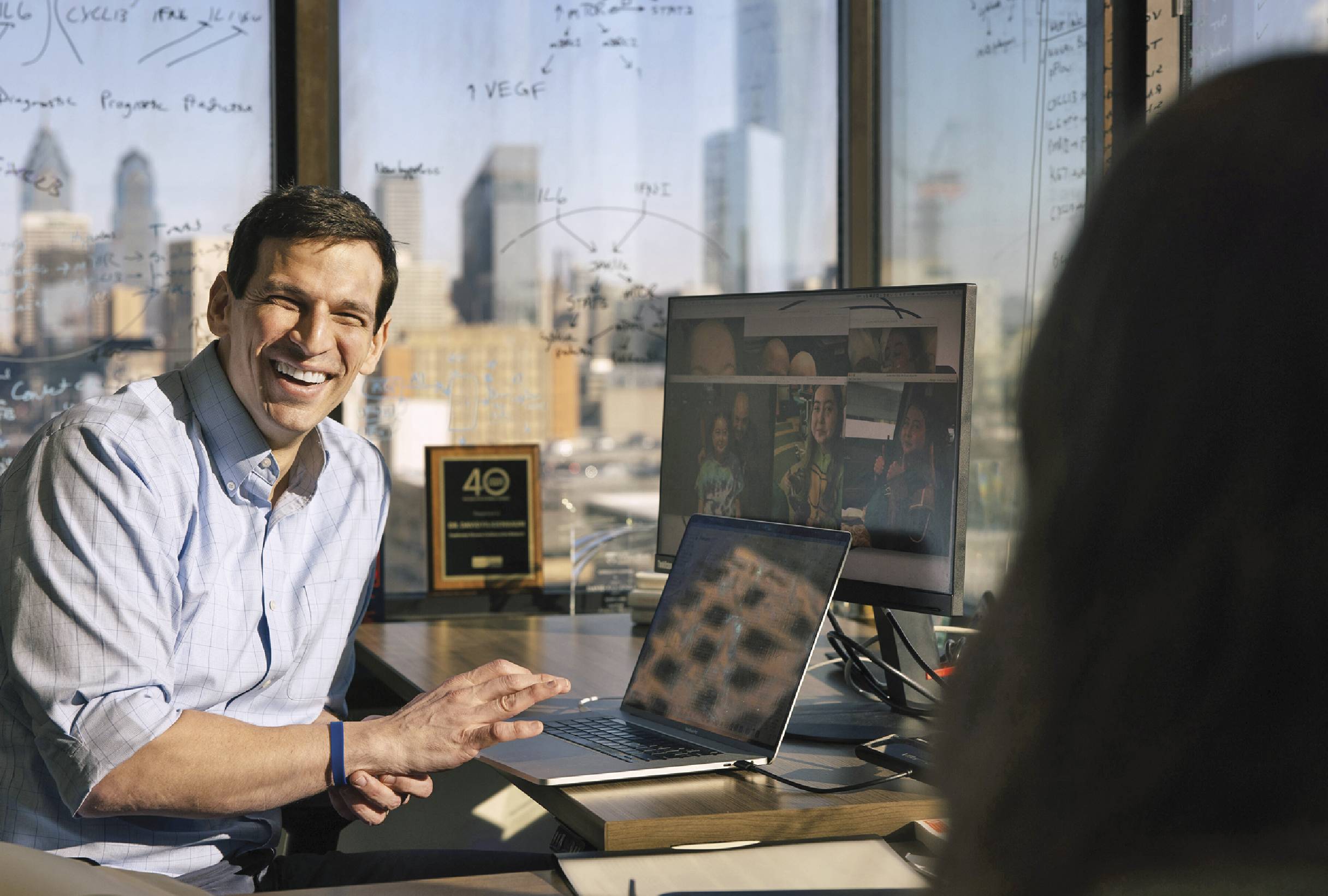 David Fajgenbaum sitting in a room with windows and equations written all over the windows. He is smiling and has multiple computer screens around him