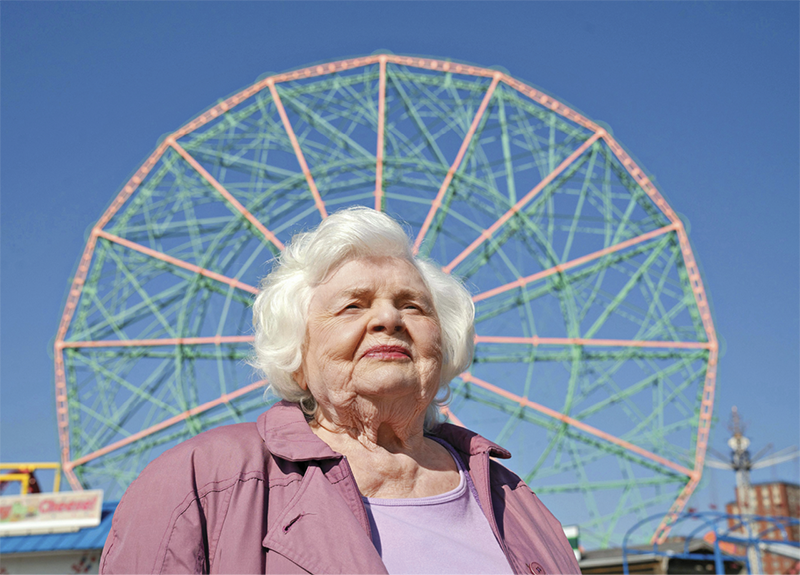 Still of June Squibb as Eleanor standing with a ferris wheel behind her