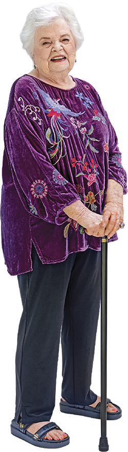 Portrait of June Squibb standing with a walking stick wearing a purple blouse and black pants