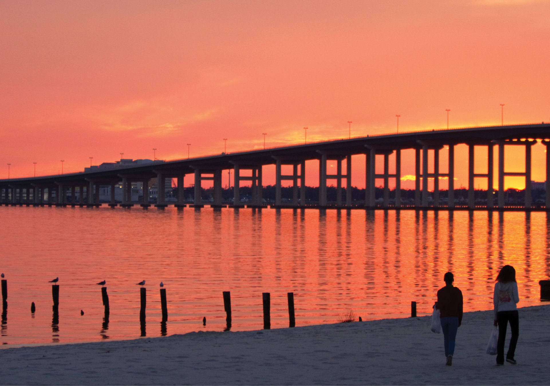 Sunset picture of Ocean Springs, Mississippi with people walking on the beach and a view of the bridge that goes over the water