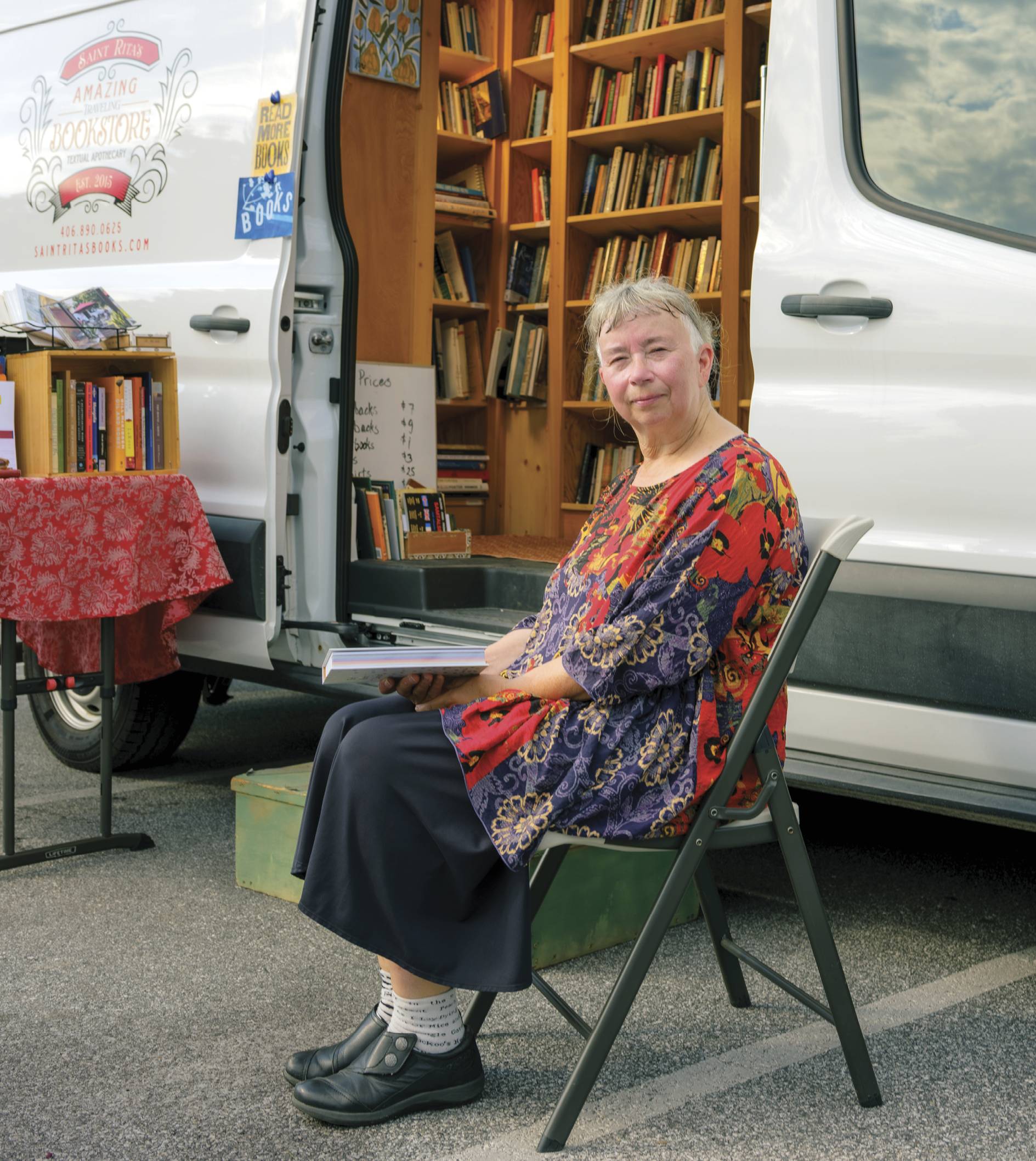 Photograph of Rita Collins sitting on a fold out chair in front of a truck with its door open, revealing bookshelf full of books inside