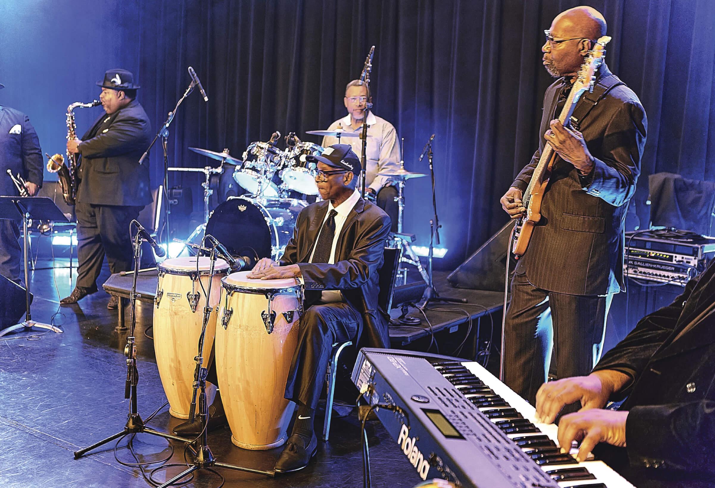 Photograph of several men on the stage playing various instruments from, the piano to conga drums, guitar and the trumpet