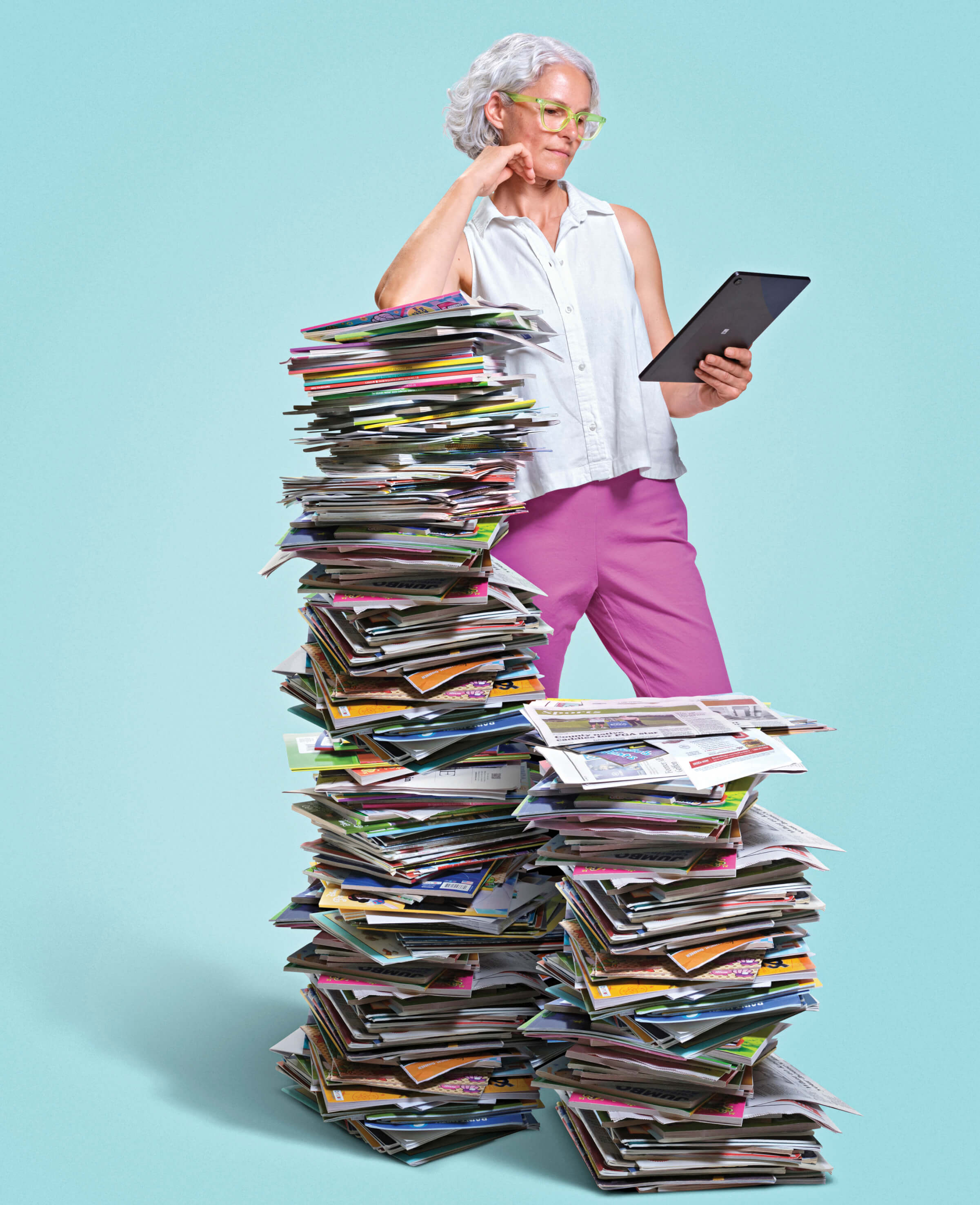 Woman leaning on a huge stack of newspapers and magazines. In her hand she is reading on a digital tablet
