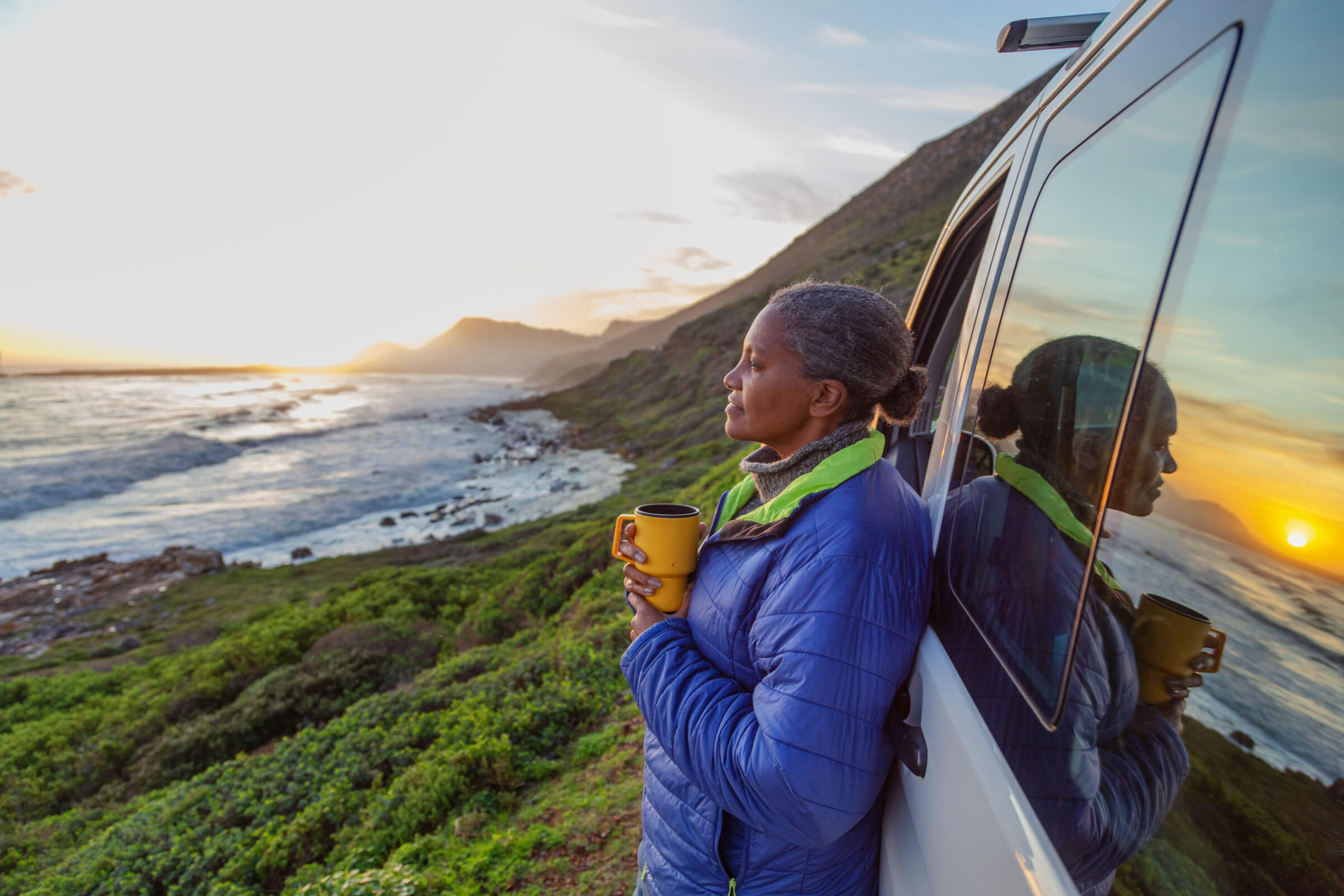 Photo of a woman with a cup of coffee, leaning against a car and looking out to the ocean