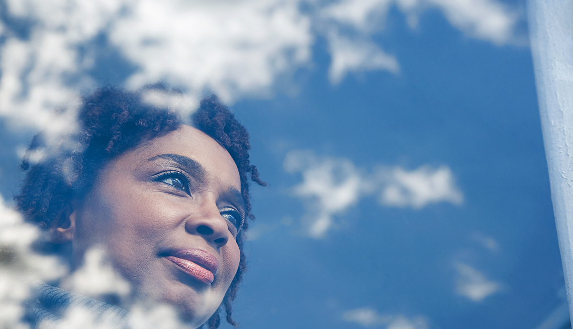 woman looks out of window at blue sky; white clouds reflect off glass around her face