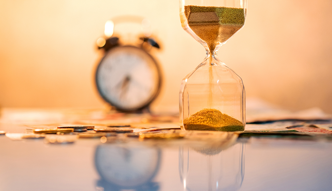 Un reloj despertador con timbres al lado de un reloj de arena y algunas monedas y billetes.