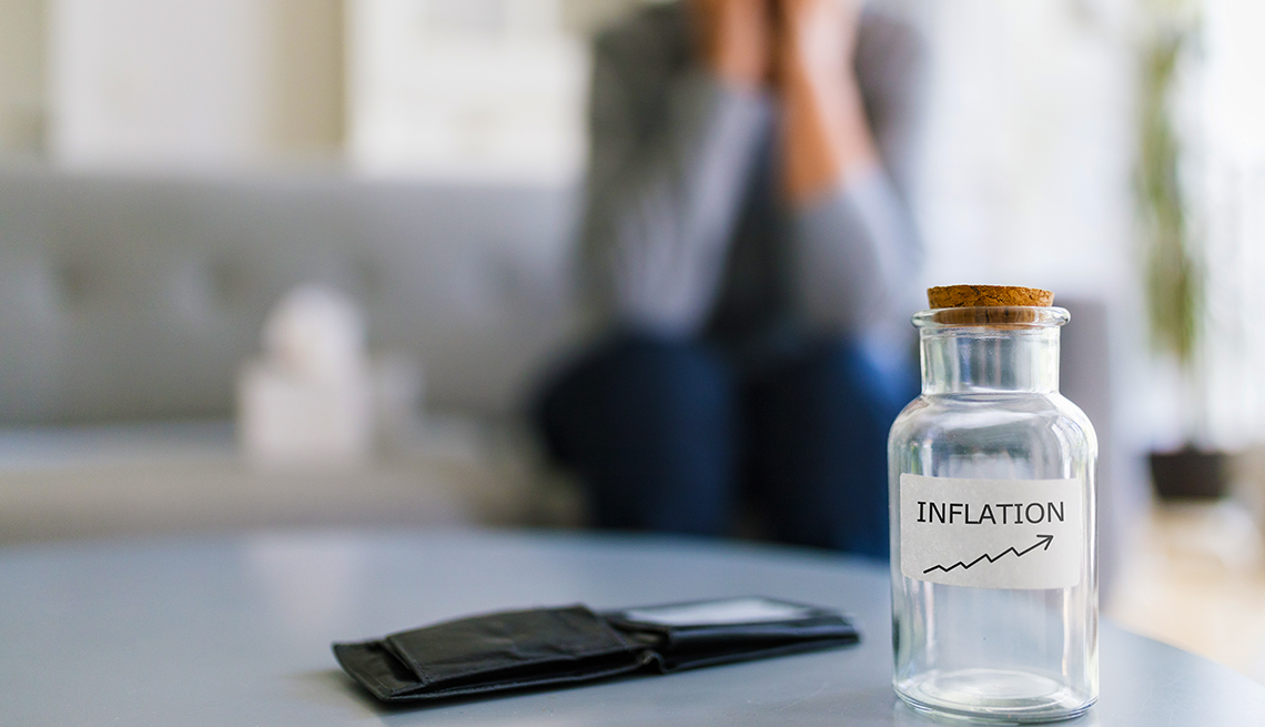 A distressed woman in soft focus sits on a sofa behind an empty wallet and empty jar labeled "Inflation".