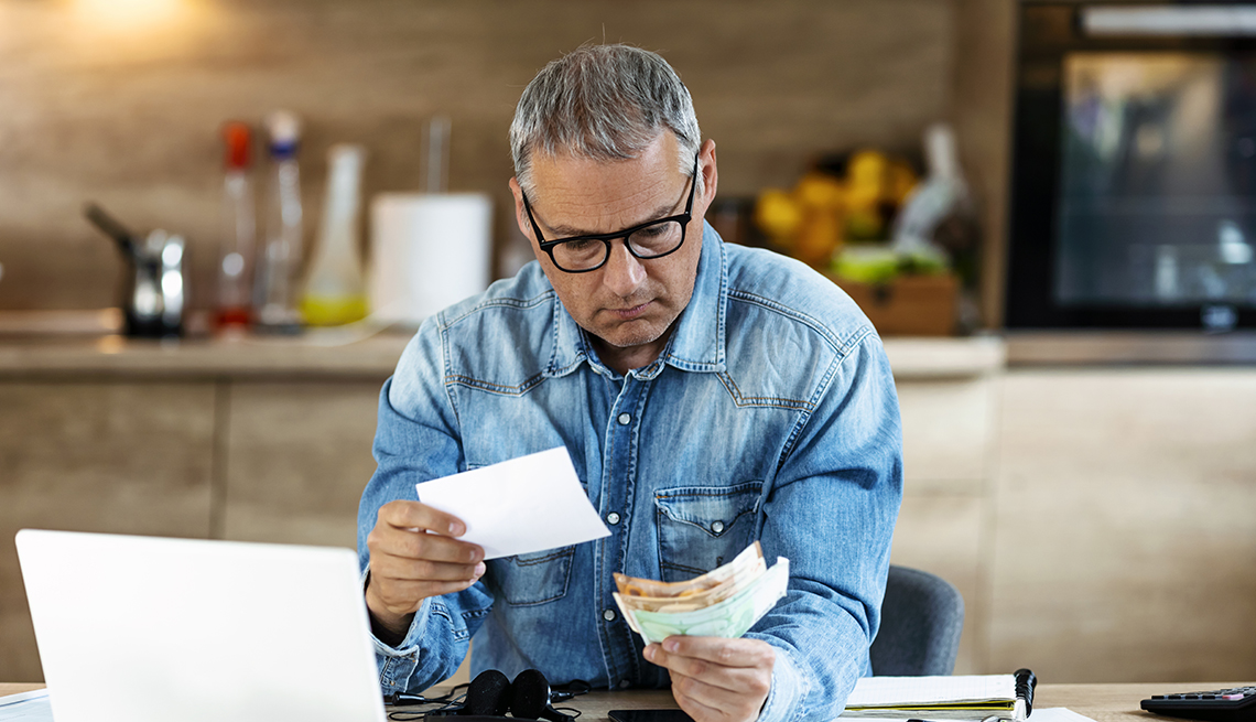 man wearing glasses with worried look on his face sits next to an open laptop computer and examines invoices and bills 