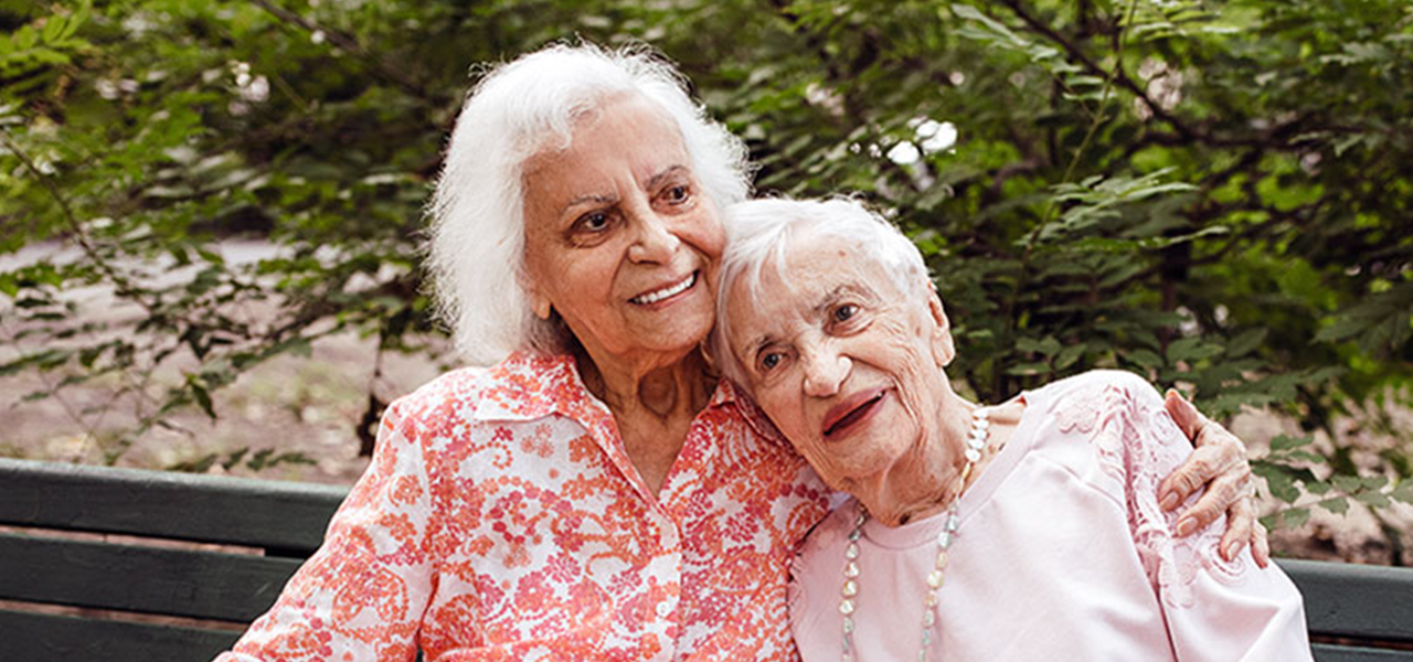 Two white haired older women hugging and laughing on a bench.