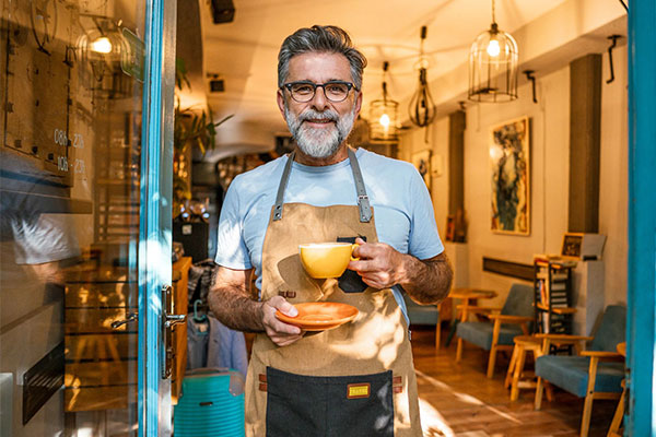 Smiling man with salt and pepper hair and beard holds coffee in front of open cafe door