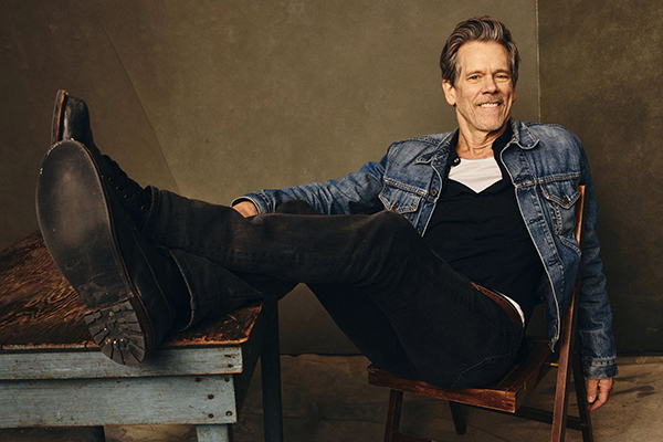 Kevin Bacon sitting in a chair with his feet up on a table.