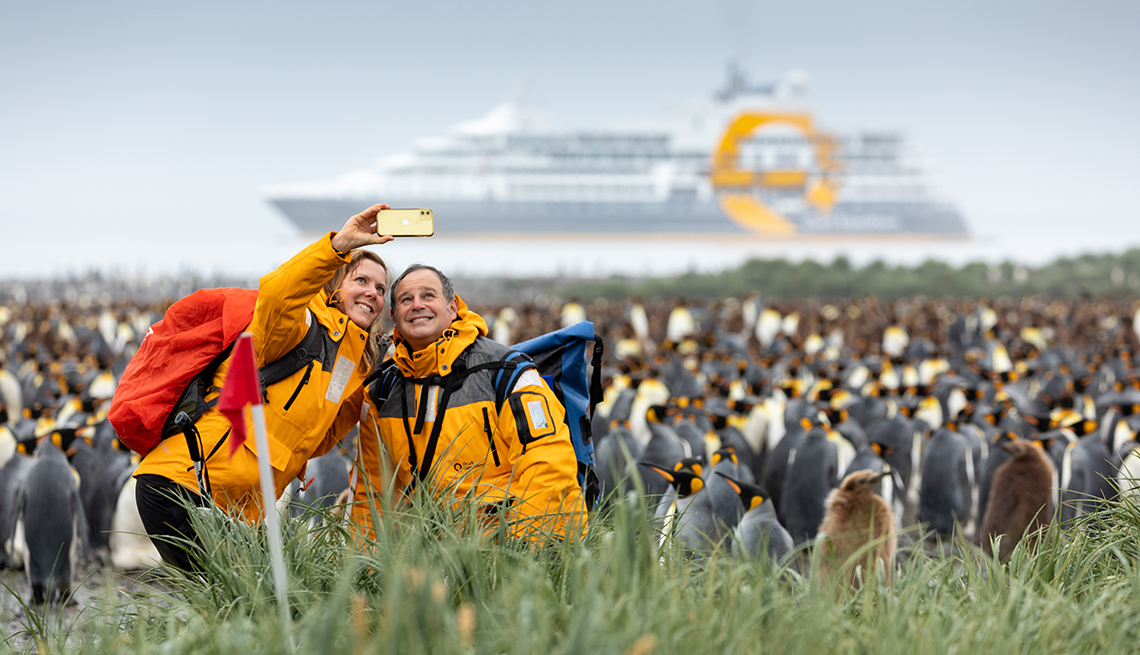 a couple taking a selfie photo while standing on shore in front of a penguin colony you can see their cruise ship in the water in the background