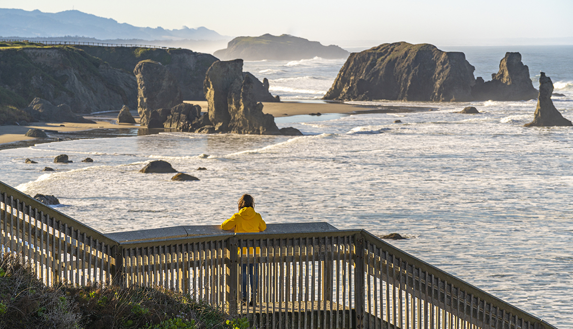 item 6 of Gallery image - a woman in a yellow jack rests her arms on a rail while looking out over a beach in oregon