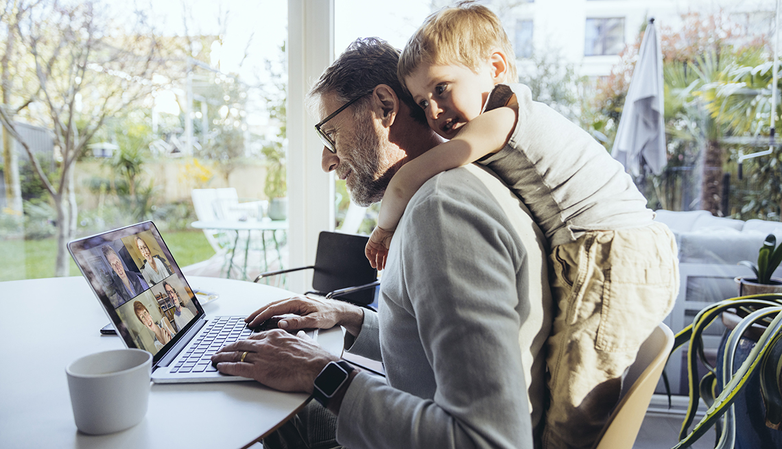 Hombre trabajando en casa frente a la computadora mientras su hijo le da un abrazo por detrás.