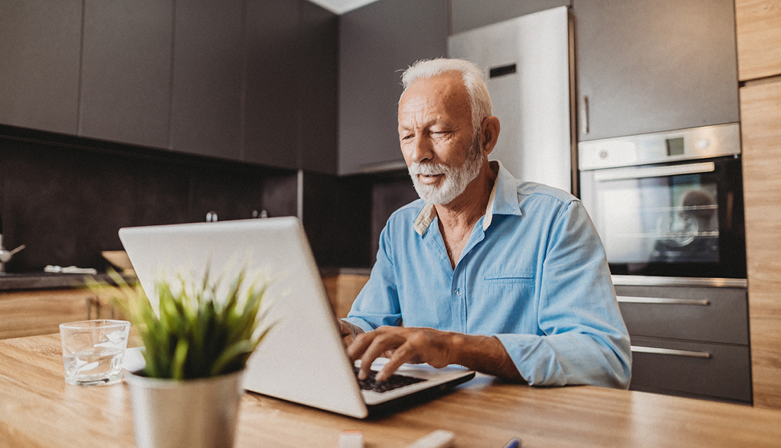 Persona mayor trabajando en su computadora desde la cocina de su casa.
