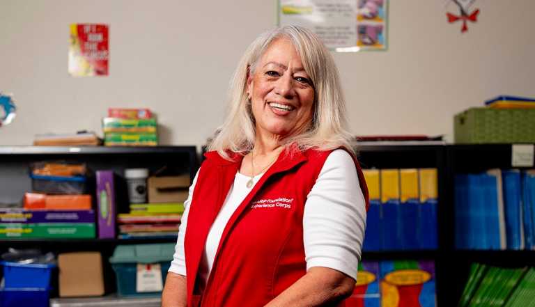 AARP Foundation Experience Corps volunteer tutor Nina Sanchez, older senior woman smiling and standing inside classroom wearing a red vest  