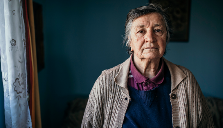 Senior woman wearing burgundy shirt and beige cardigan sweater inside her home looking at camera