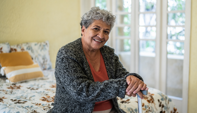 a smiling woman sitting on the edge of a bed holding a walking cane