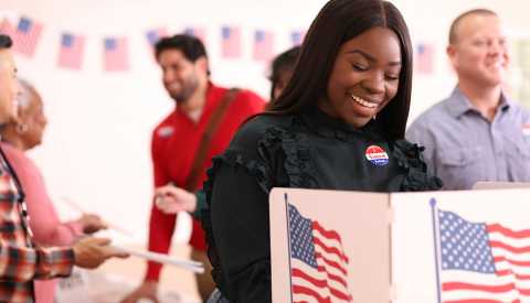 African descent, young or mid-adult woman votes in the USA election.  She stands at voting booth in polling station.   Other voters and election day registration seen in background.