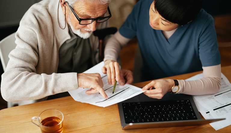 High angle view of a Japanese female caregiver doing home finance online on a computer together with her worried elderly patient at his home.