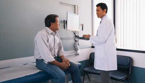 Male doctor working on computer while discussing with senior male patient in hospital.