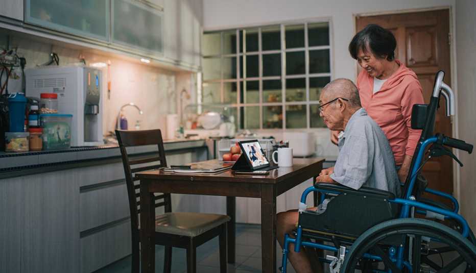 Asian senior man with wheelchair and his wife videocall his family using digital tablet in kitchen at night after dinner leisure time together.