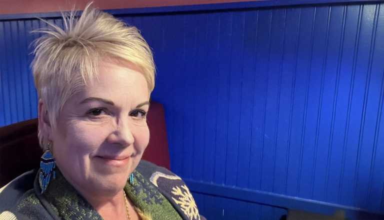 Kat Willis , senior woman blonde hair wearing blue earrings and a sweater sitting in a chair inside