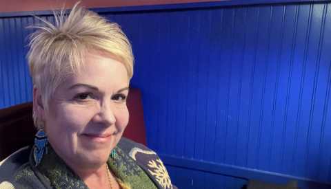 Kat Willis , senior woman blonde hair wearing blue earrings and a sweater sitting in a chair inside