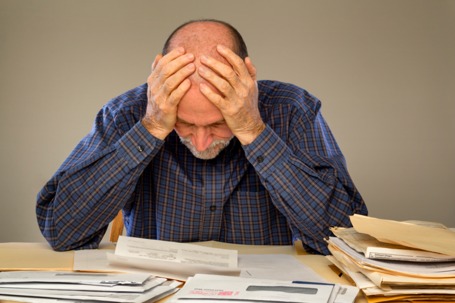 A senior adult man sitting at a table or desk stacked with papers and envelopes looking down with his hands on his head. 