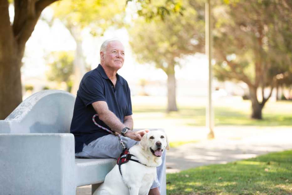 A white, older veteran sits with his dog in a park on a sunny day