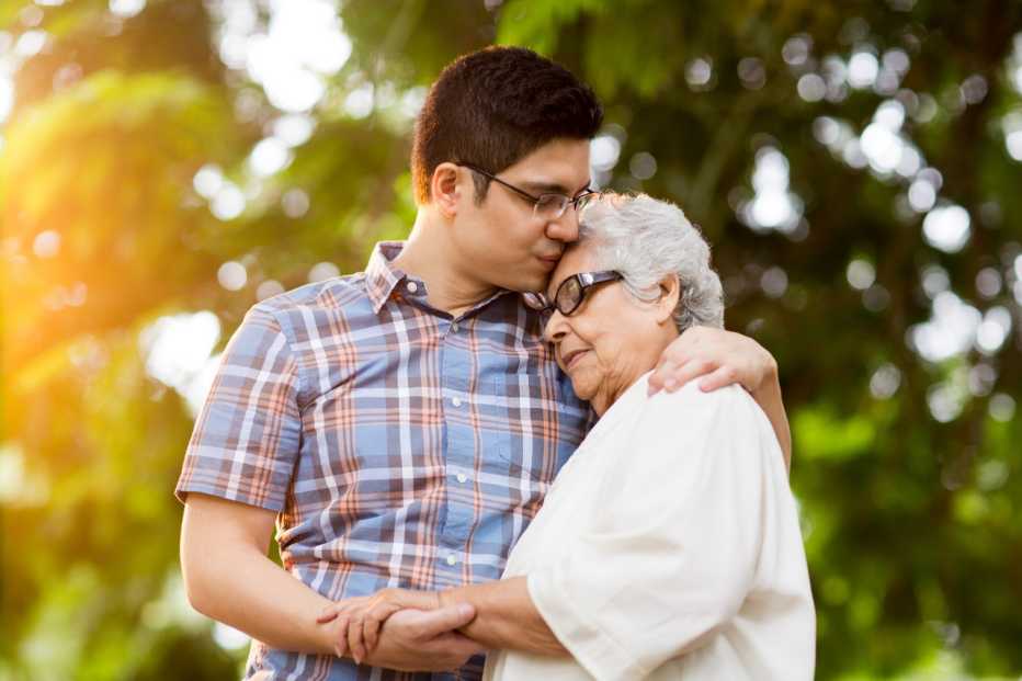 A latin adult grandson and a senior grandmother embracing with eyes closed in a horizontal waist up shot outdoors.