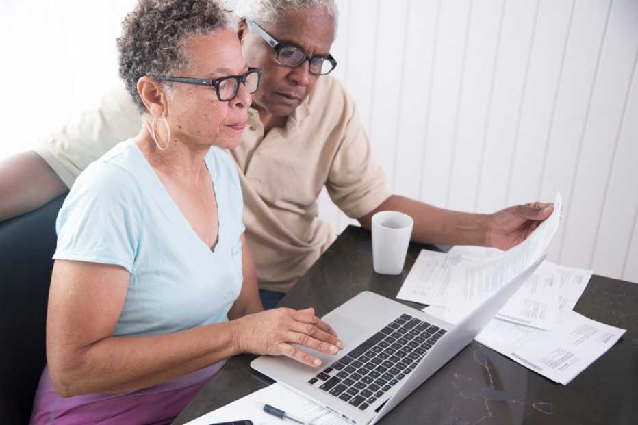 Senior couple sitting at table, using laptop, looking at paperwork