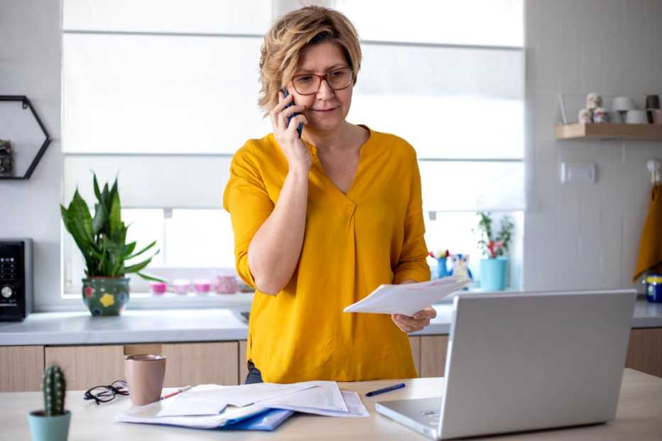 Concentrated mature woman calculating finance, money, using laptop computer and smart phone at home workplace table, counting budget, paying bills, taxes, rent, mortgage fees