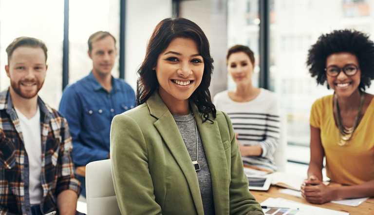 A woman sitting and smiling in a boardroom meeting with her colleagues behind her