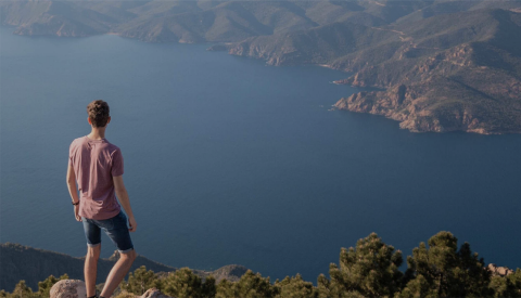 A man standing on the top of a peak looking out at the landscape and body of water