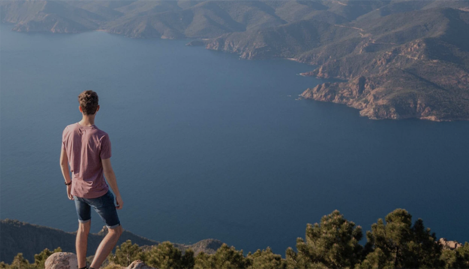 A man standing on the top of a peak looking out at the landscape and body of water