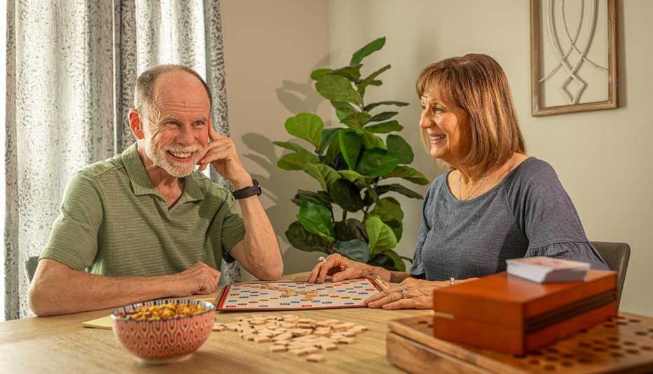 Two adults playing a word board game at a dining table with letter tiles and snacks in a cozy home setting.